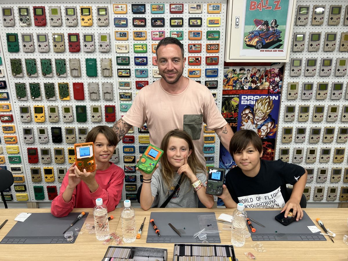 A dad and three kids proudly holding up the Game Boys they just built, in front of the cartridge wall